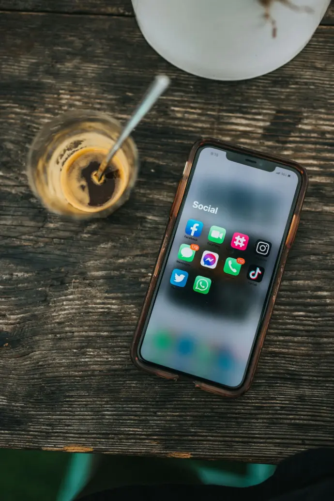 mobile phone sitting on a wood table with social media tab open beside a cup of glass of coffee with a spoon in it