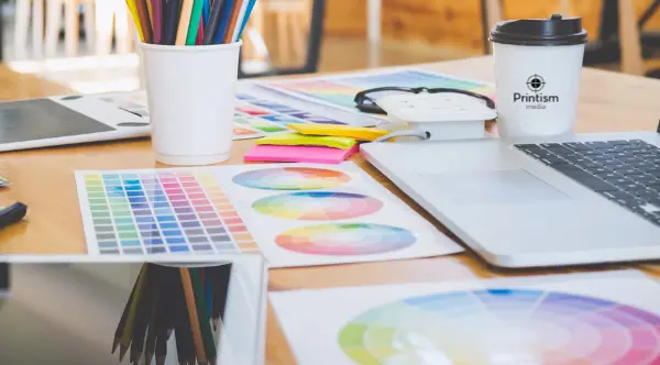 sheets of paper with colour charts on a table with a pot of pens and a laptop